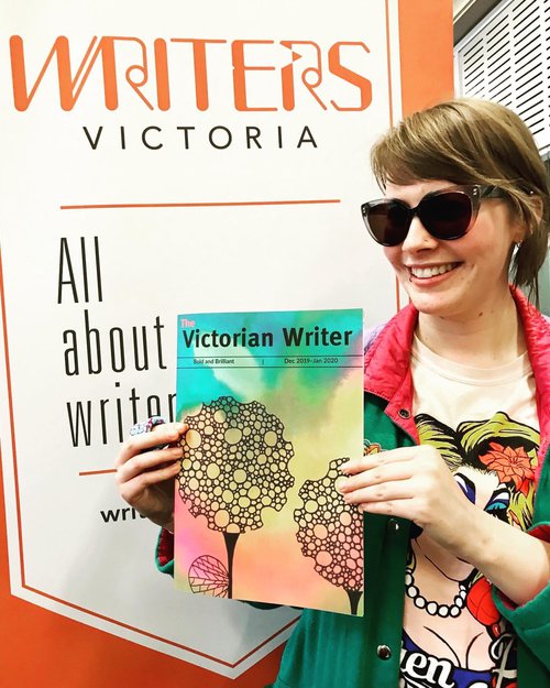 Alex standing in front of a poster for Writers Victoria and holding a copy of The Victorian Writer publication